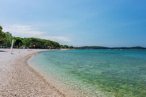 A pebble beach with clear turquoise water, a flag, and trees in the background. - Apartments Petra (Fazana)