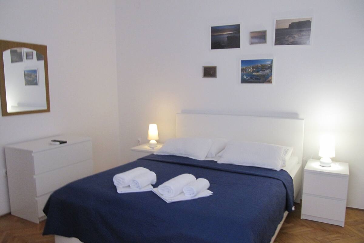 A hotel room with a bed, two bedside tables, a nightstand, a dresser, and a wall with framed pictures.