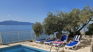 A swimming pool with clear blue water, surrounded by lounge chairs and a view of the sea and mountains. - Rooms Falcon (Gradac)