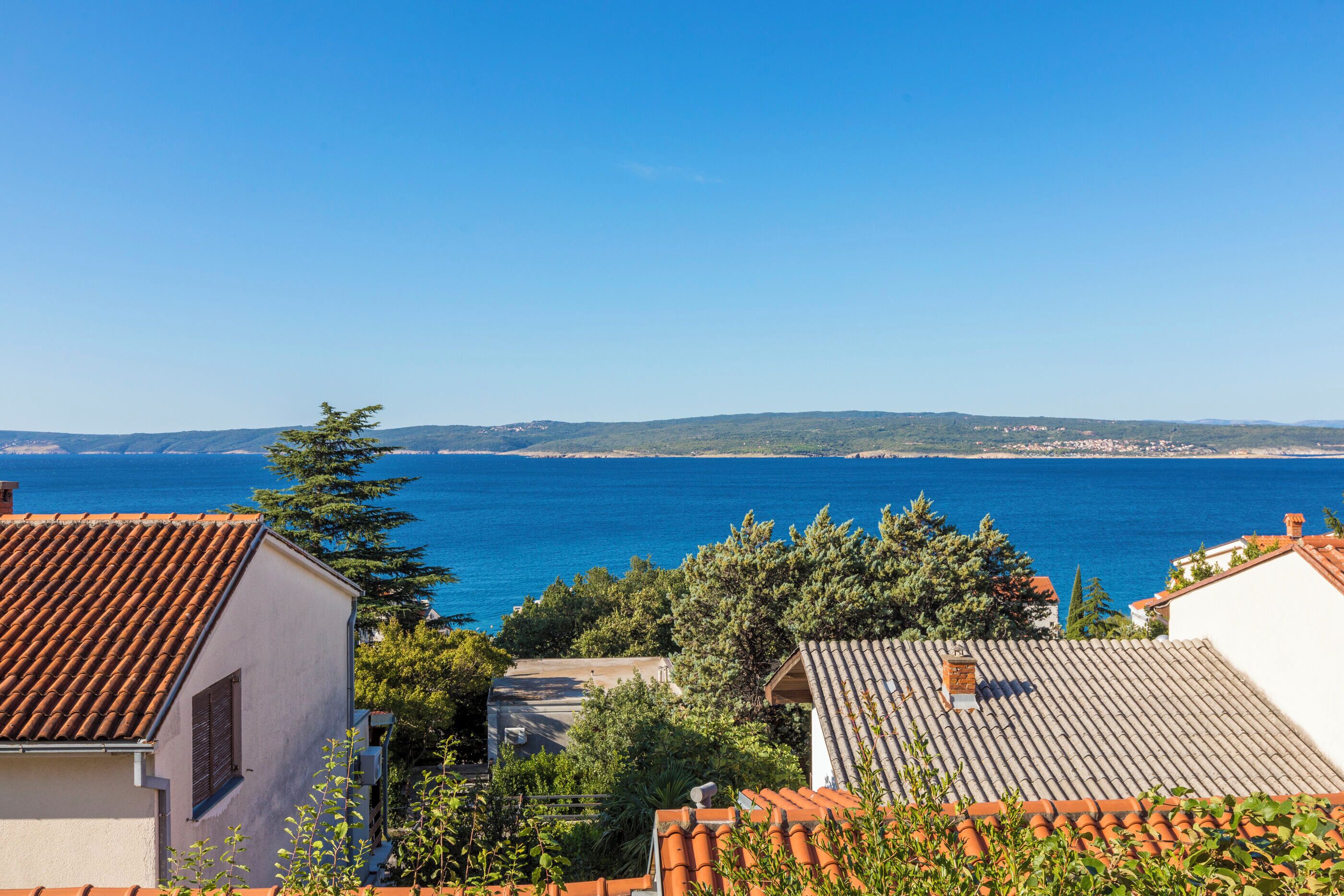 Vista sulla costa con edifici dalle tegole color terracotta, un mare blu limpido e colline in lontananza.