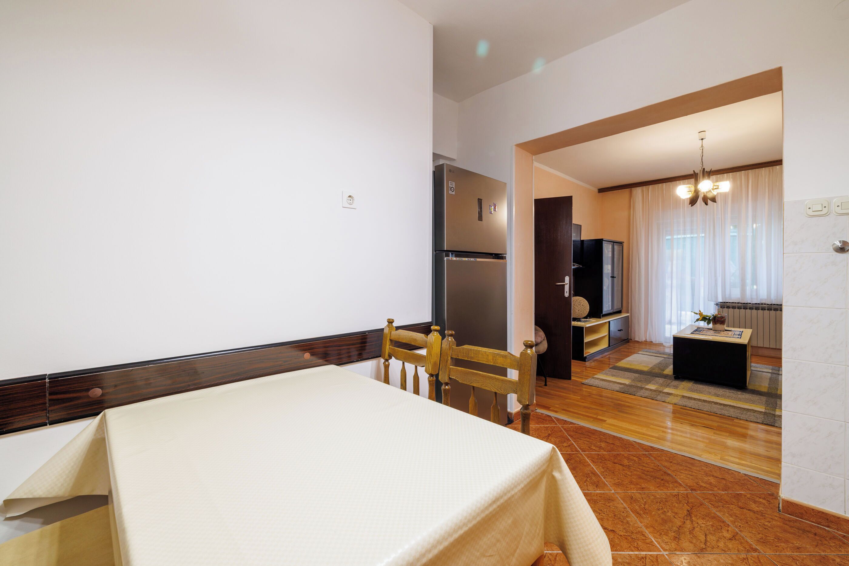 A hotel room with a bed, a wooden chair, a dining table, a refrigerator, a television, and a window with curtains.