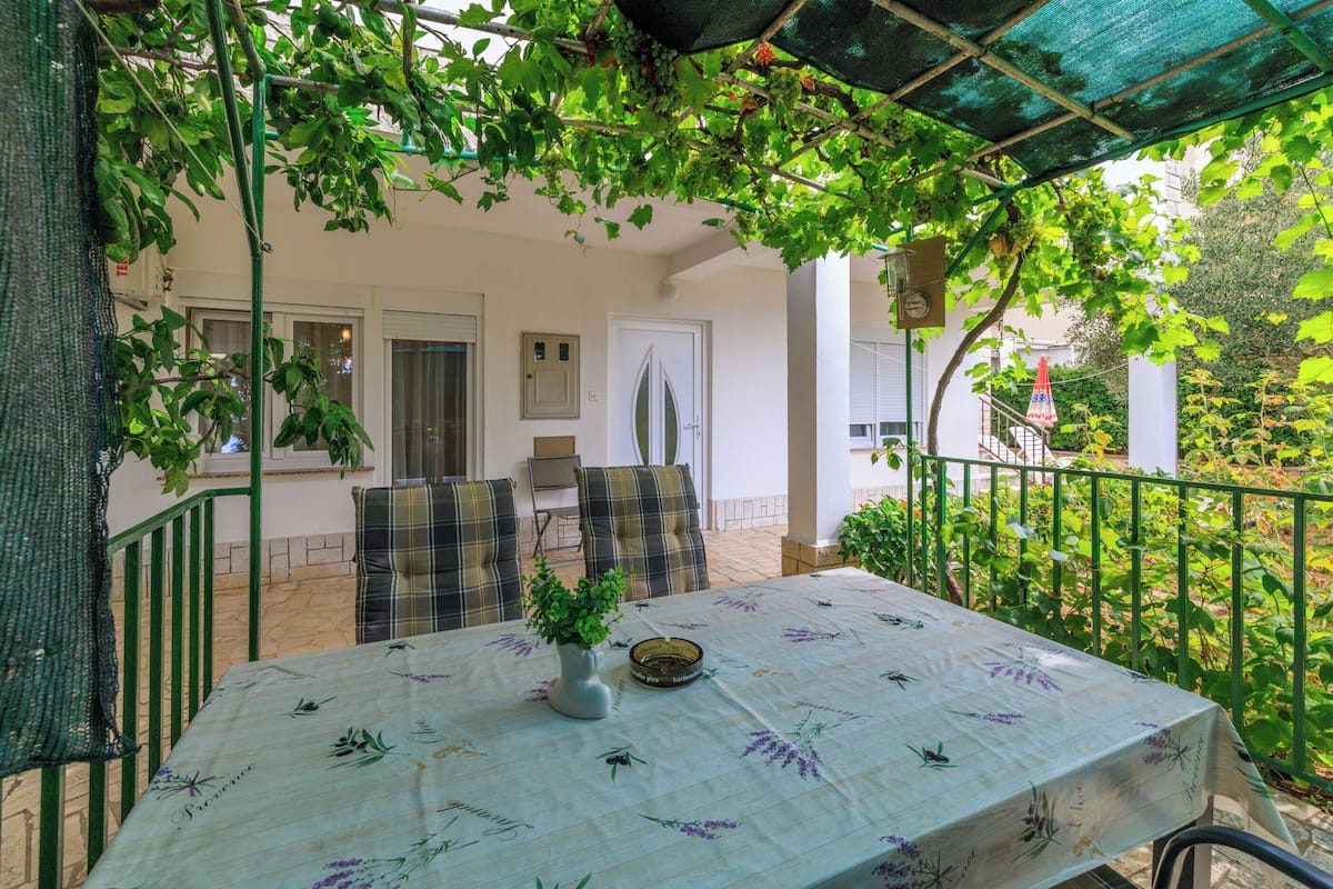 Une terrasse extérieure avec une table recouverte d’une nappe à motifs floraux, un vase avec des plantes vertes et une rambarde en métal.