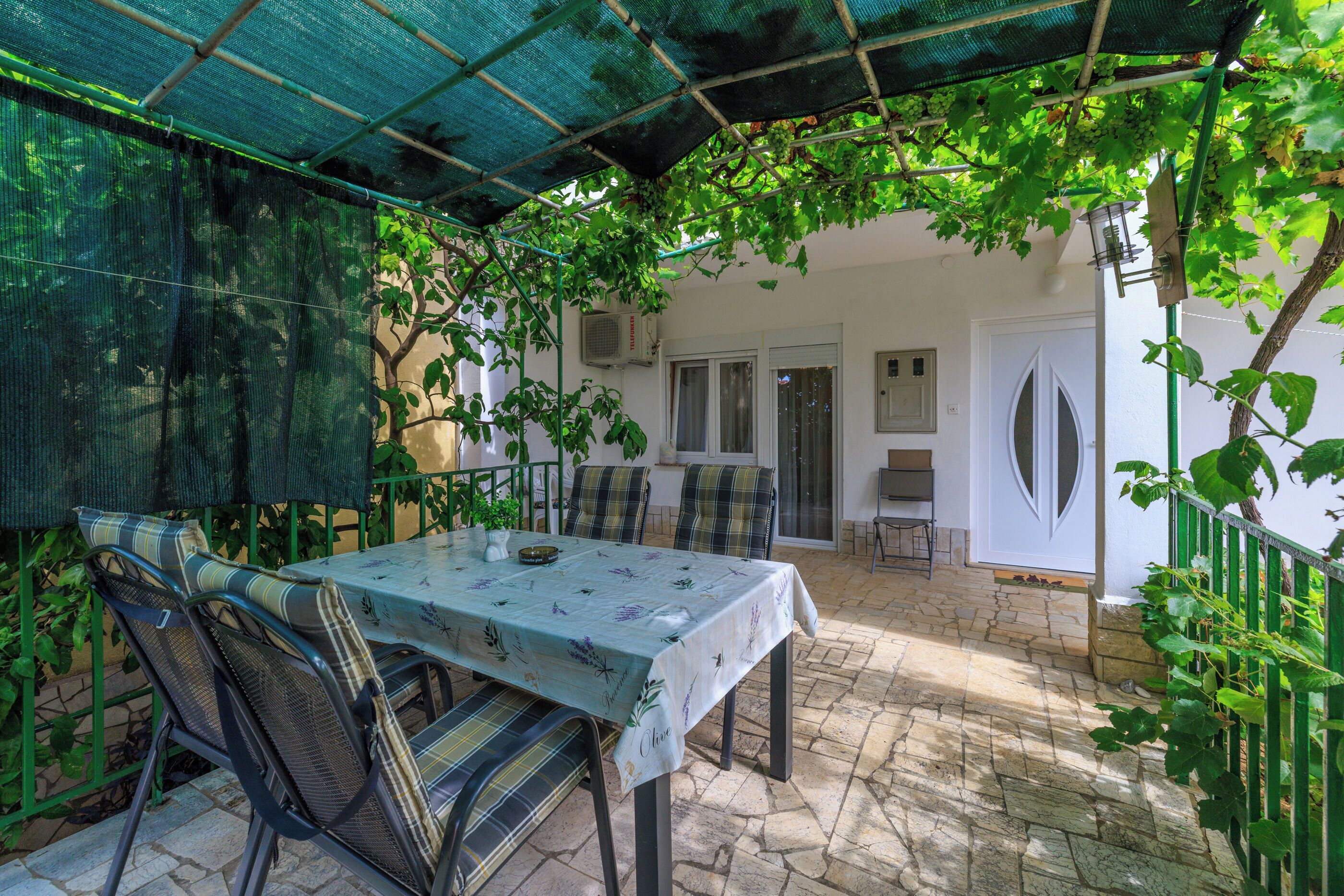 An outdoor patio with a table and chairs, covered by a green canopy, and surrounded by greenery.