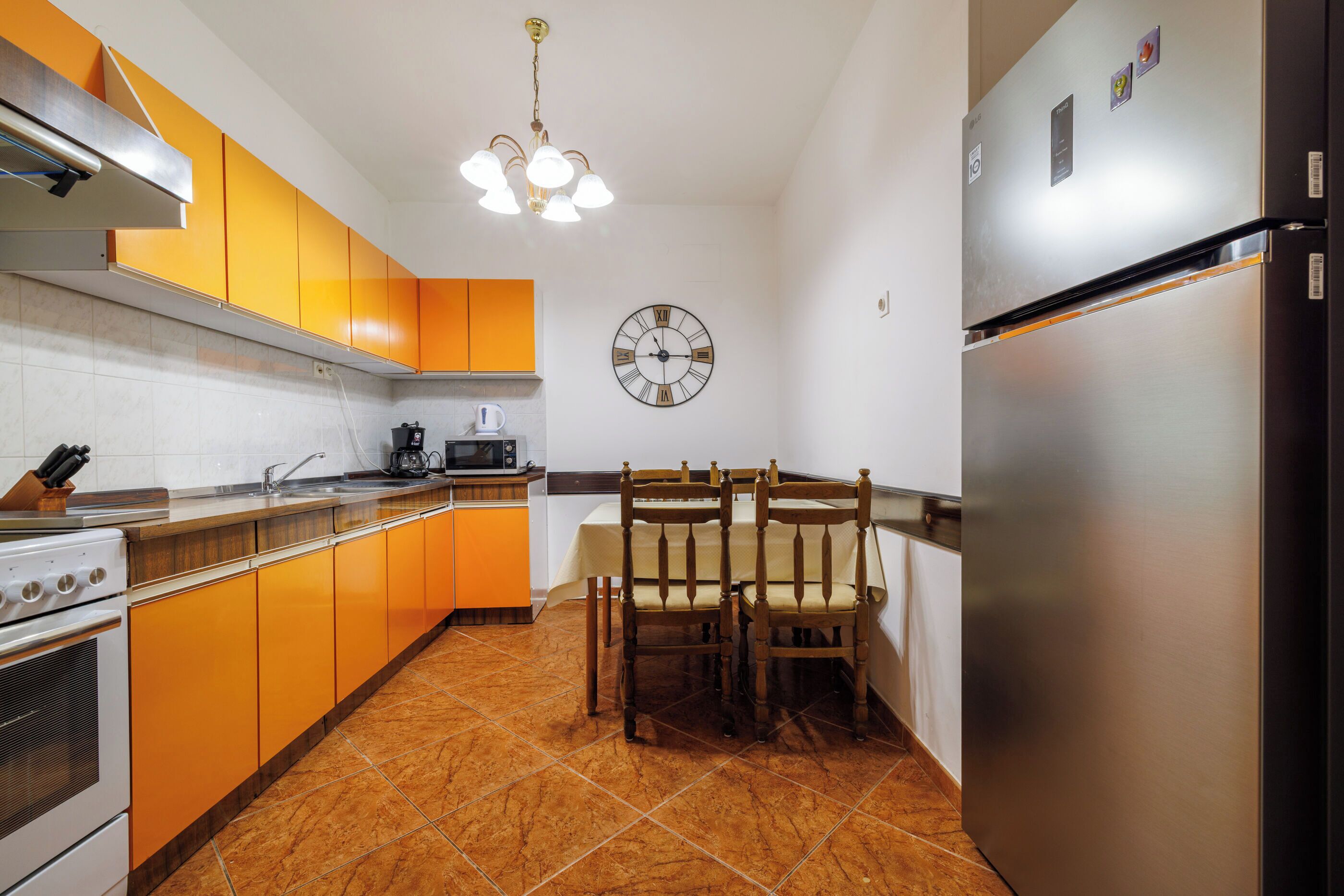 A kitchen with orange cabinets, a stainless steel refrigerator, a wooden dining table with chairs, a clock on the wall, and a tiled floor.