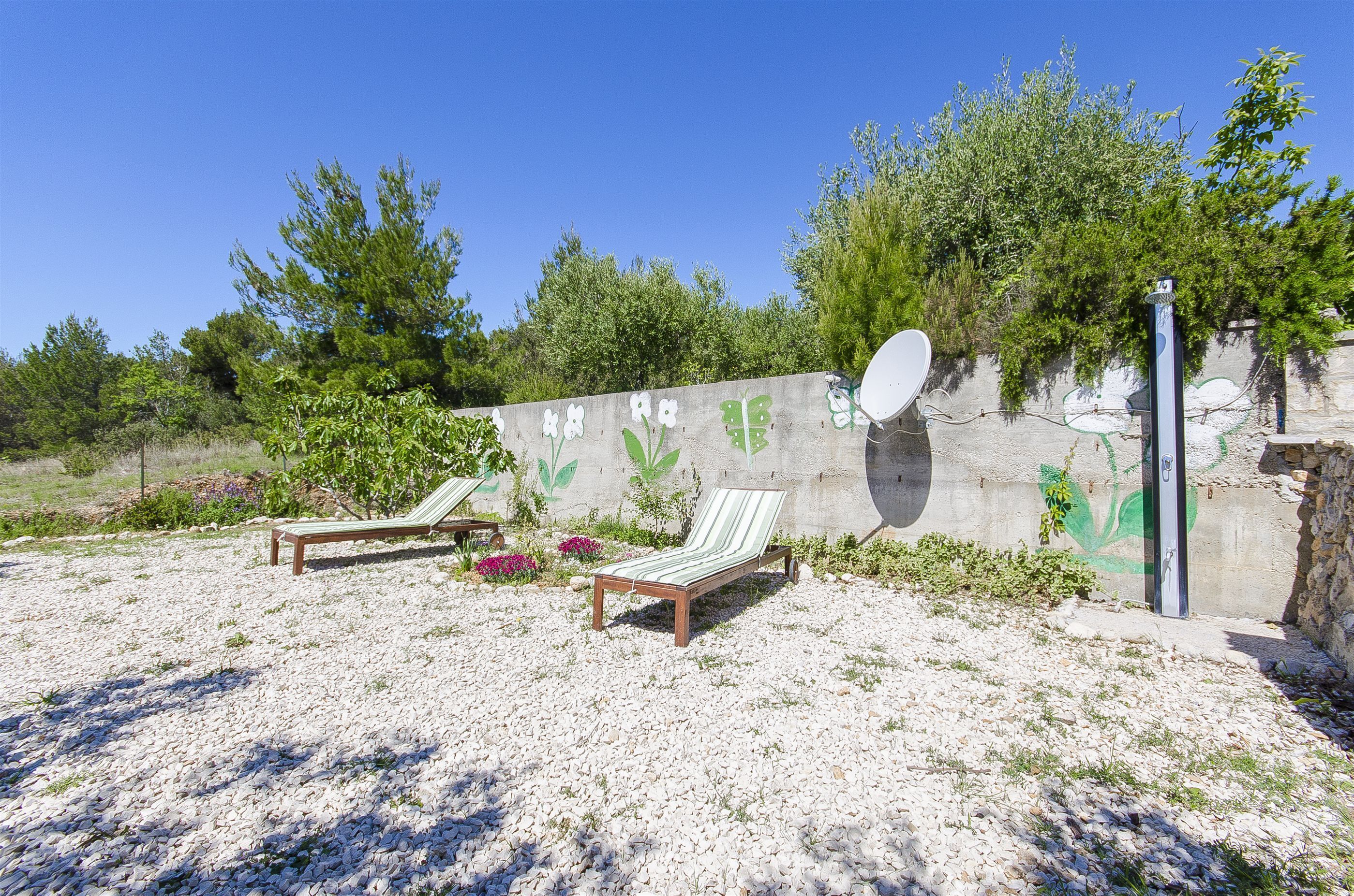 A beach area with two wooden lounge chairs, a pebble-covered ground, and a wall with green leafy patterns.