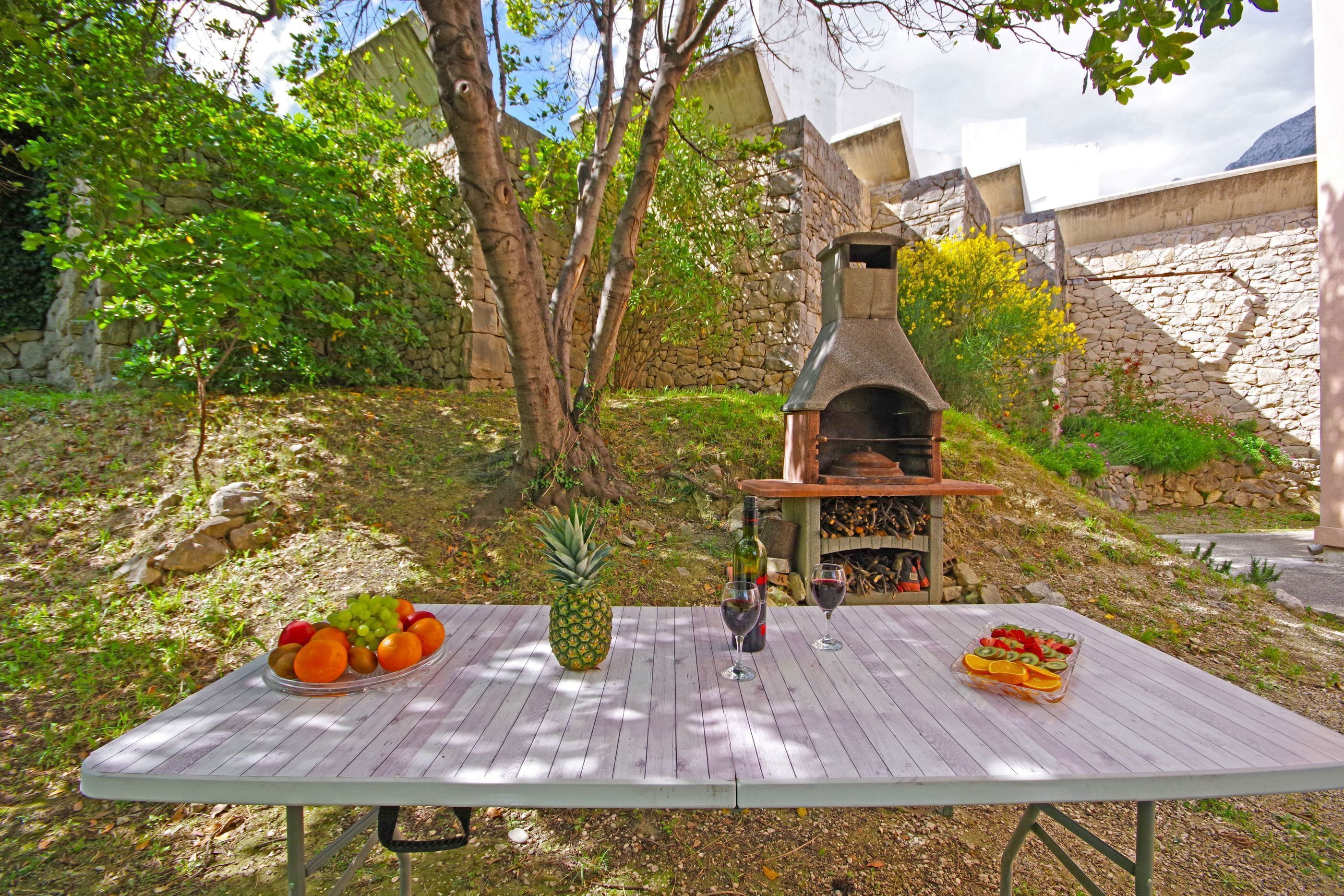 An outdoor dining area with a stone wall, a pizza oven, and a table set with fruits and wine glasses.