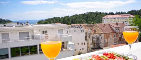 A balcony with a fruit platter and two glasses of orange juice, overlooking a cityscape with buildings and a view of the sea.