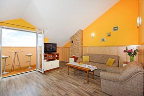 Een moderne woonkamer met een bank, salontafel en een televisie. Er is een balkon met een glazen deur en een klein tafeltje met twee krukjes.