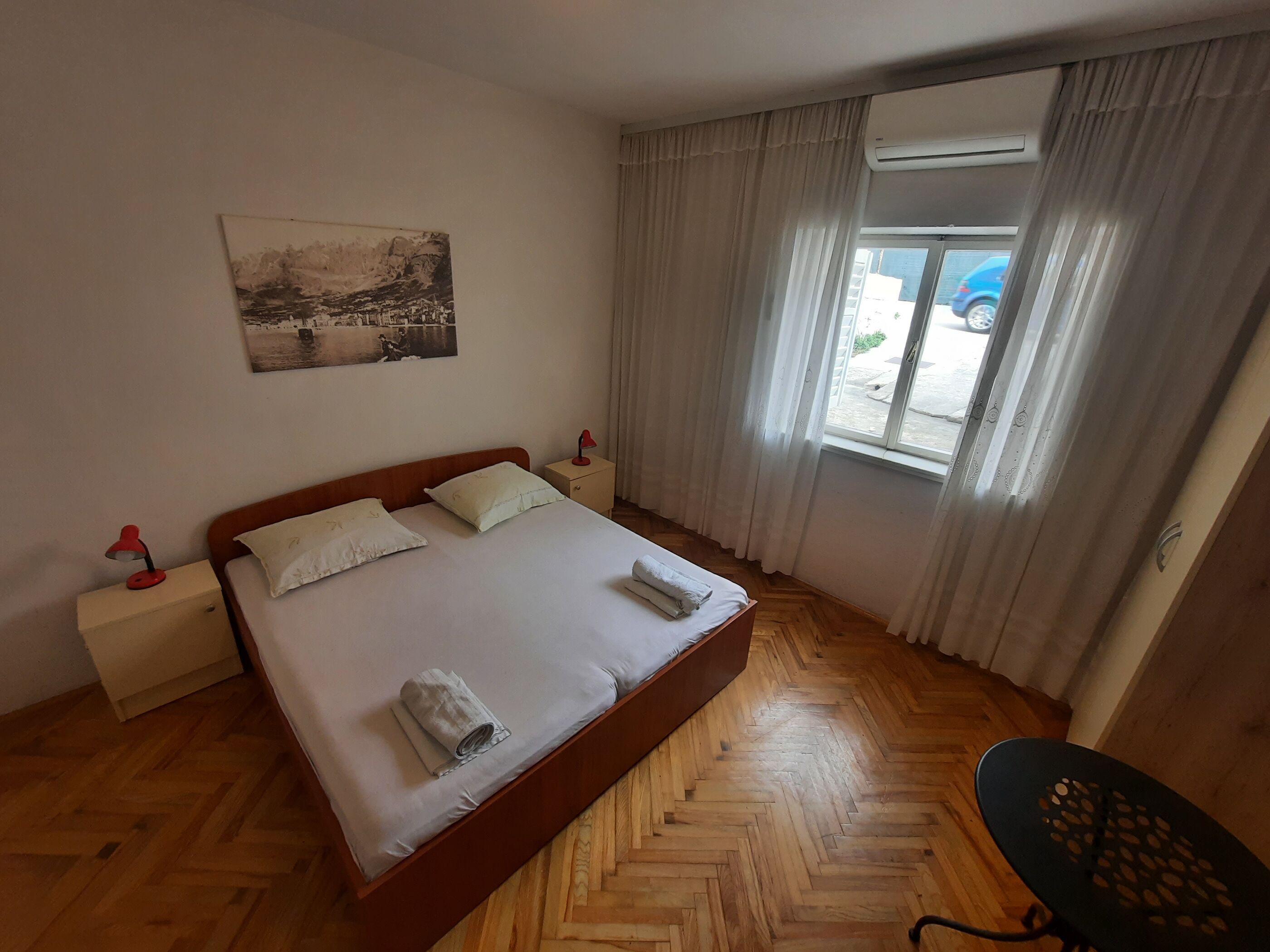 A bedroom with a wooden herringbone floor, a bed with white linens, a nightstand with a lamp, a window with curtains, and a painting on the wall.
