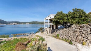 Una propiedad costera con un muro de piedra, un camino que lleva a un edificio blanco con balcón, y vista al mar.