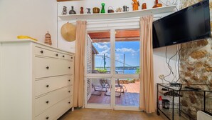 A room with a sliding glass door leading to an outdoor area with a view of a bridge and a stone wall. There is a TV mounted on the wall, a white cabinet with drawers, and a cart with various items. - House Bubble (Crikvenica)