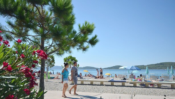 Ein Strand mit Menschen, die spazieren gehen und sitzen, einem klaren Himmel und einem Gewässer im Hintergrund.