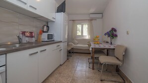 Una cucina compatta con mobili bianchi, un forno a microonde e un lavandino. C'è un'area pranzo con tavolo e sedie, e una finestra con tende.