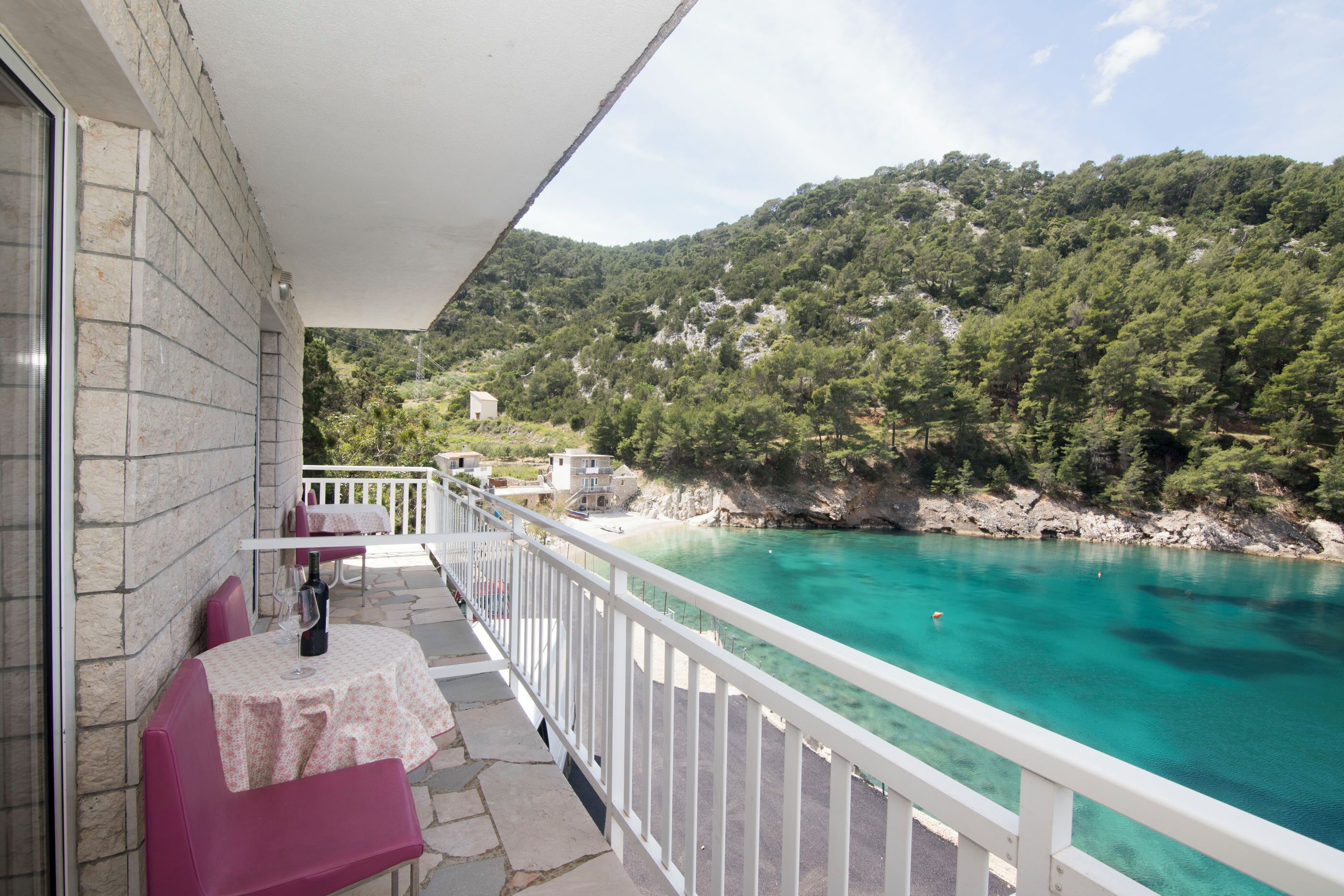 Un balcón con mesa y sillas, con vista al mar turquesa y un paisaje montañoso.