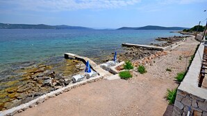 A coastal area with clear water, rocky shore, and a paved walkway with umbrellas. - House Merica (Trogir)
