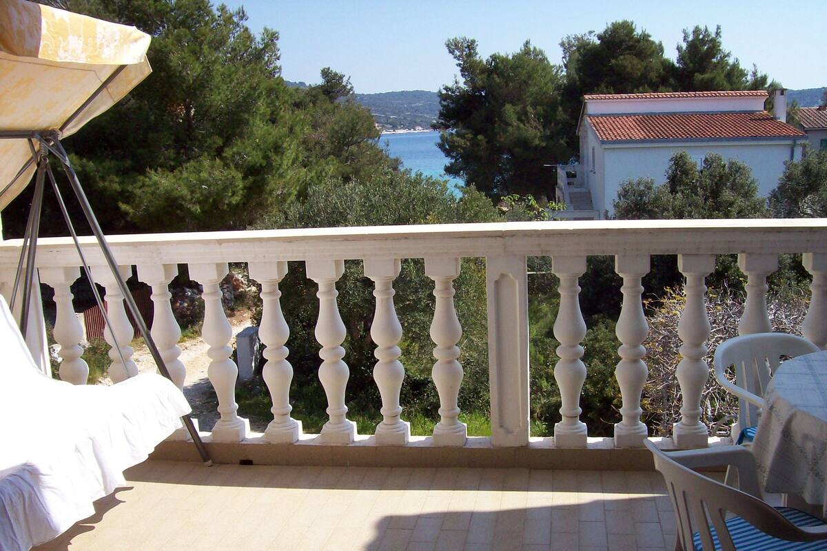 Un balcon avec une balustrade blanche, une table avec une chaise, et une vue sur la mer et les arbres.