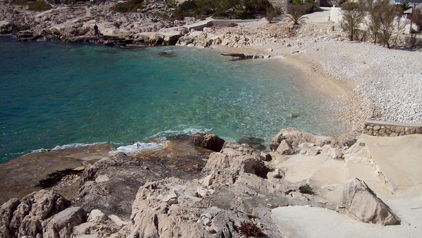 A rocky beach with clear turquoise water, a sandy area, and buildings in the background. - Apartments Barac (Marina)