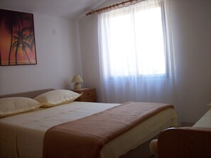 Ein Schlafzimmer mit Bett, Nachttisch, Lampe und einem Fenster mit durchsichtigen Vorhängen.