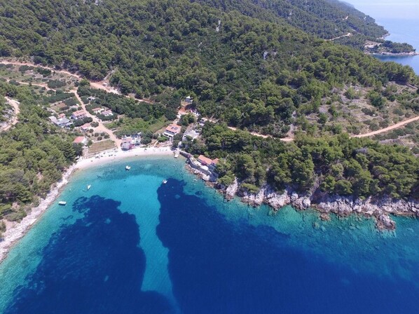 Une vue aérienne d’une zone côtière avec une plage, des eaux d’un bleu limpide et quelques bâtiments dispersés.
