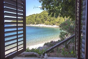A coastal view through a wooden door with a clear blue sea, a sandy beach, and a lush green hill in the background. - Apartments Zeljko (Jelsa)