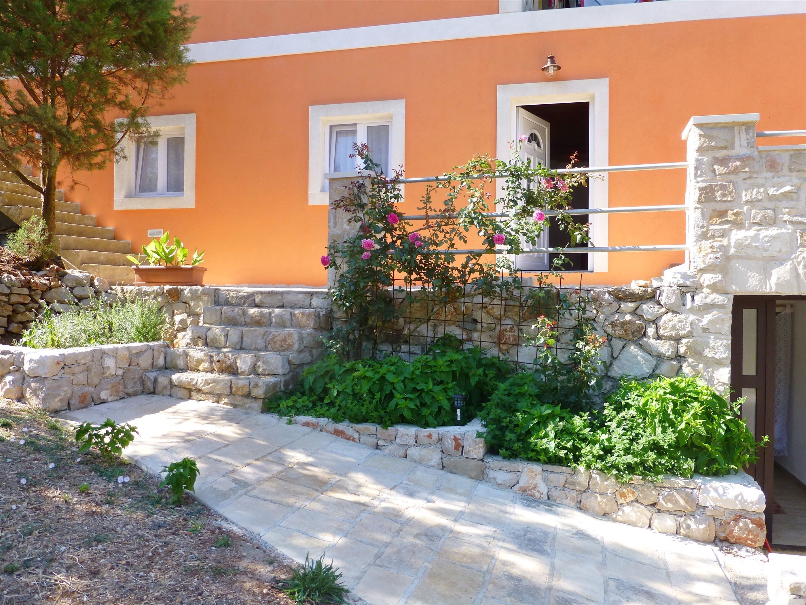 A stone pathway leading to a building with orange walls and white trim. There are potted plants and a small tree in the foreground.