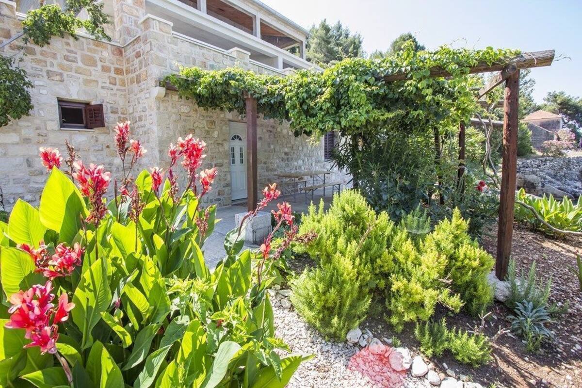 A stone building with a garden featuring various plants and a wooden pergola.