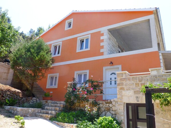 A two-story building with a stone staircase leading to the entrance, surrounded by greenery and a clear sky. - Apartments Zeljko (Jelsa)