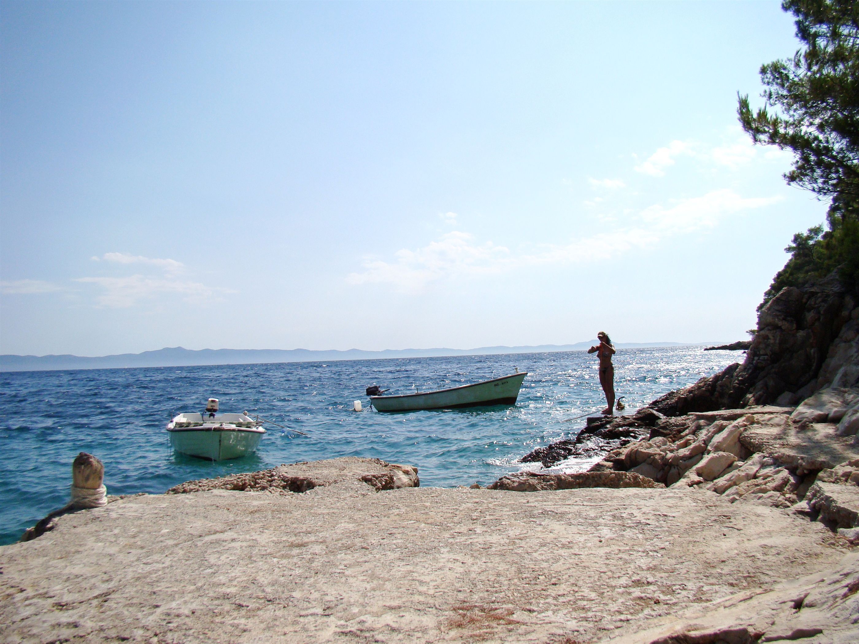 A rocky shoreline with a clear view of the sea, a small boat, and a person standing on the rocks.