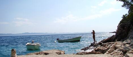 A rocky shoreline with a clear view of the sea, a small boat, and a person standing on the rocks.