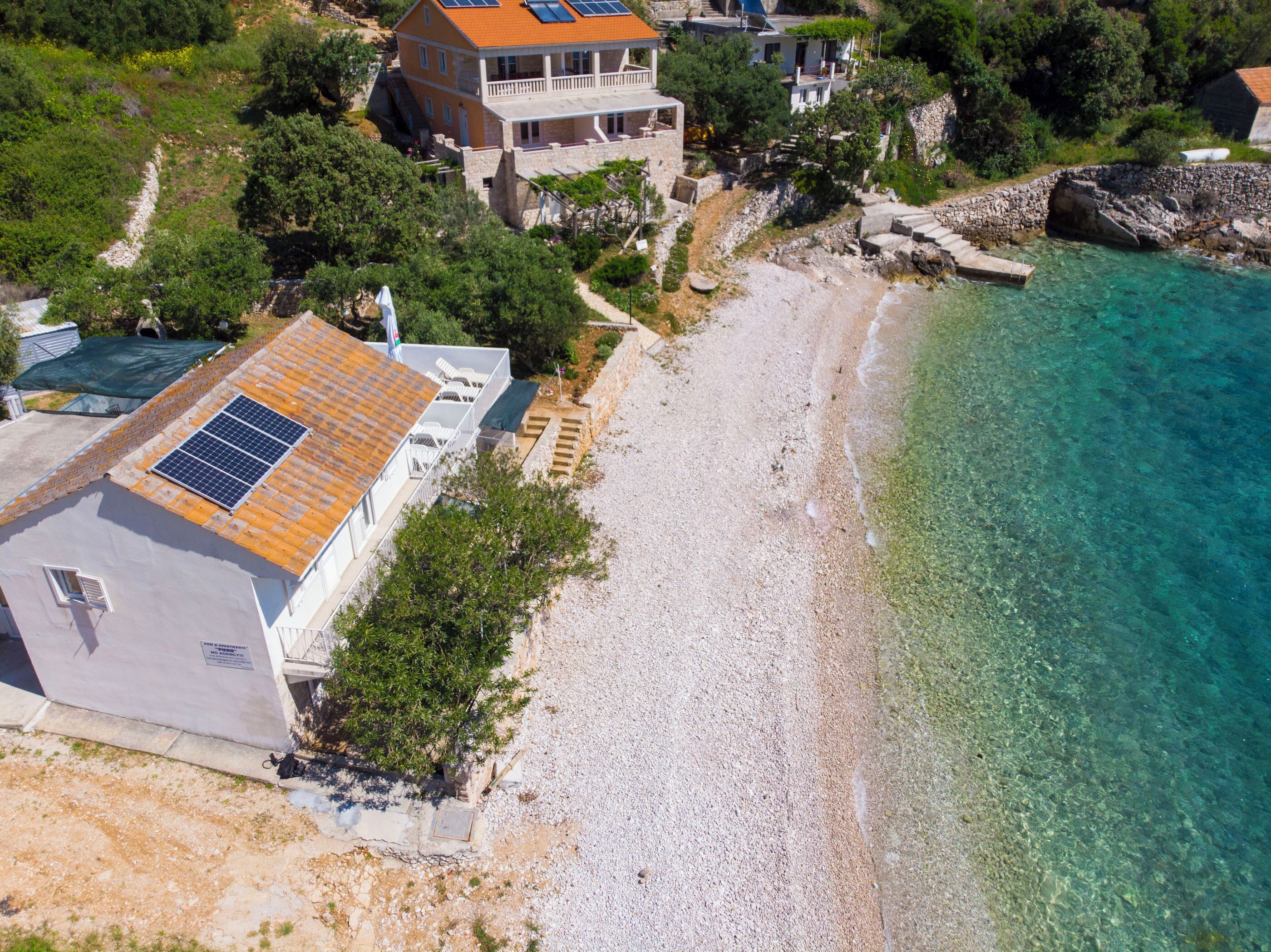 A coastal property with a large house featuring a solar panel roof, a beach, and clear waters.
