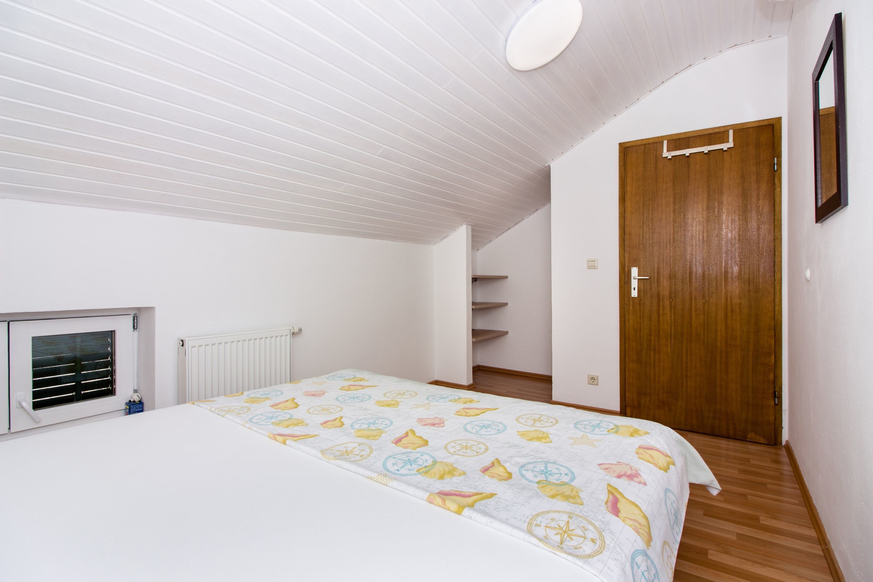 Un dormitorio con cama, ventana con persianas, radiador, puerta de madera y escalera.