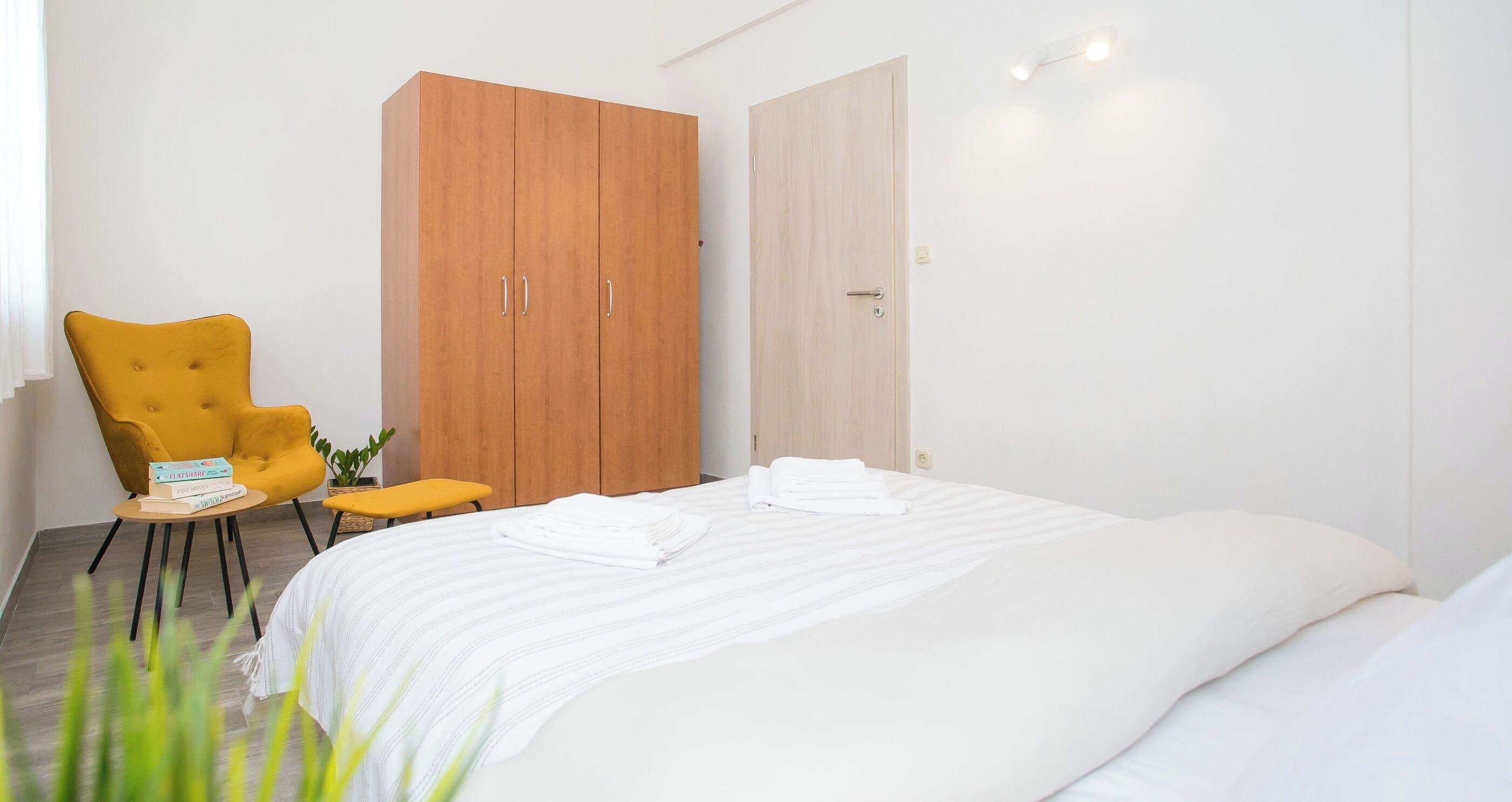 A bedroom with a bed, a wardrobe, a yellow armchair, and a small table with books.