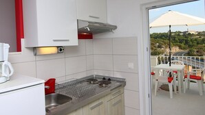 A compact kitchen with a built-in stove, a sink, and a white refrigerator. There is a red accent wall and a view of a balcony with outdoor seating. - Apartments Huljev (Primosten)