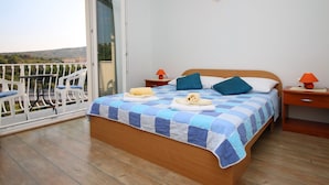 A bedroom with a bed, two bedside tables with lamps, a balcony with chairs, and a view of the landscape. - Apartments Huljev (Primosten)