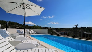 A swimming pool with white lounge chairs and umbrellas, surrounded by a fence and hills in the background. - Apartments Huljev (Primosten)