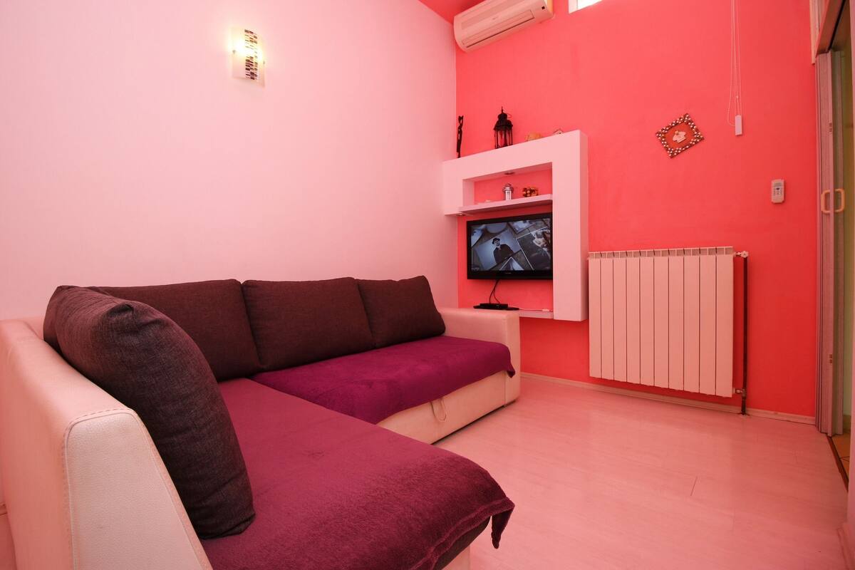 Una stanza dalle pareti rosa con un divano bianco, un televisore su una mensola e un termosifone.