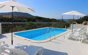 Ein Swimmingpool mit zwei weißen Sonnenschirmen, umgeben von weißen Liegestühlen und einem Metallzaun.