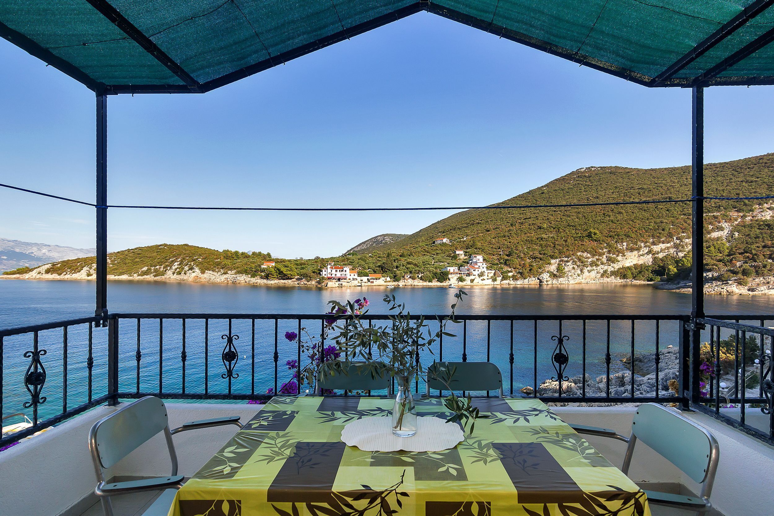 A covered outdoor seating area with a table and chairs, overlooking a body of water and a hilly landscape.
