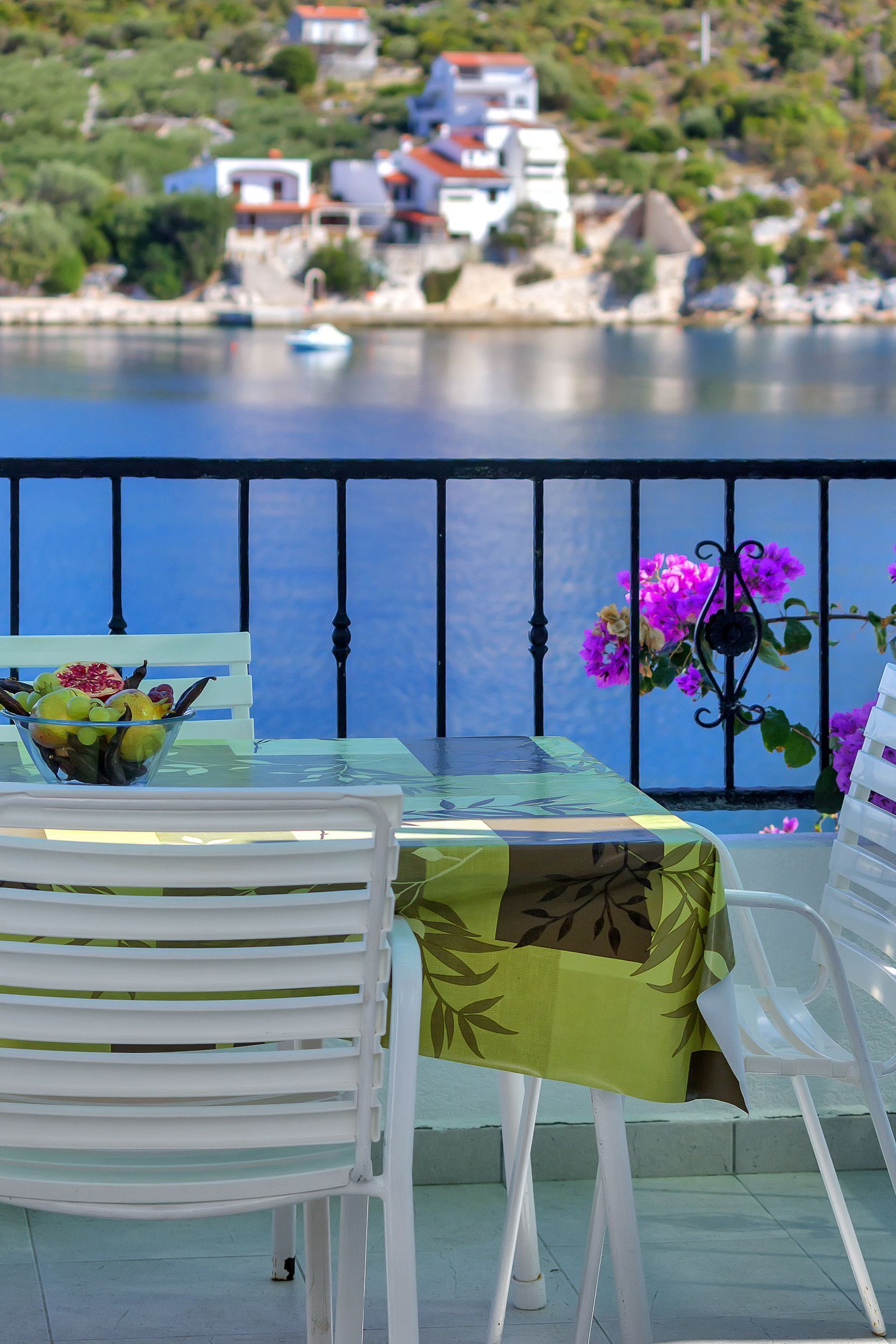 A table with a glass top and a green tablecloth, a white chair, and a balcony with a view of a lake and houses in the background.