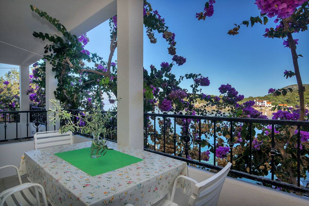 A balcony with a table set for two, surrounded by purple bougainvillea flowers, overlooking a body of water and a hillside.