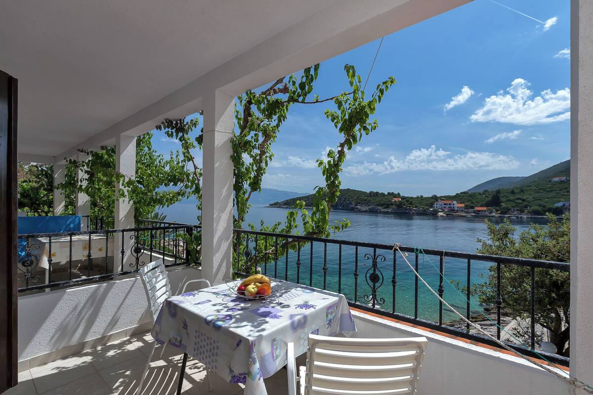 A balcony with a table set for dining, overlooking a body of water and a distant coastline.