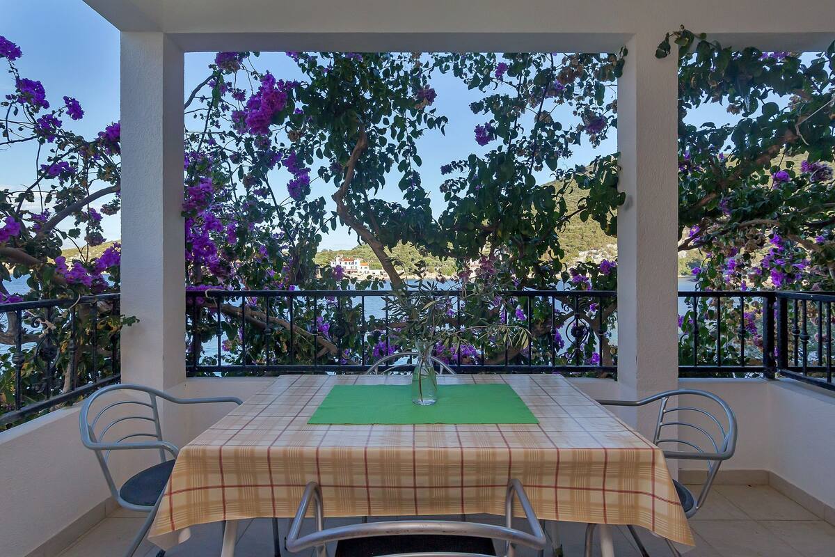A balcony with a table and chairs, covered by a checkered tablecloth, surrounded by purple flowers and a view of the sea.