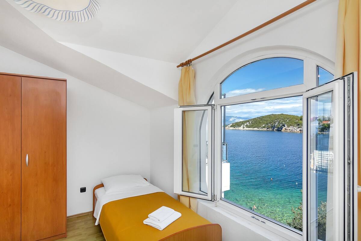 A bedroom with a bed, a wooden wardrobe, and a window with a view of a body of water and a hilly landscape.