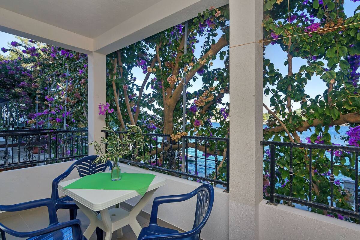 A balcony with a table and chairs, surrounded by lush greenery and purple flowers.