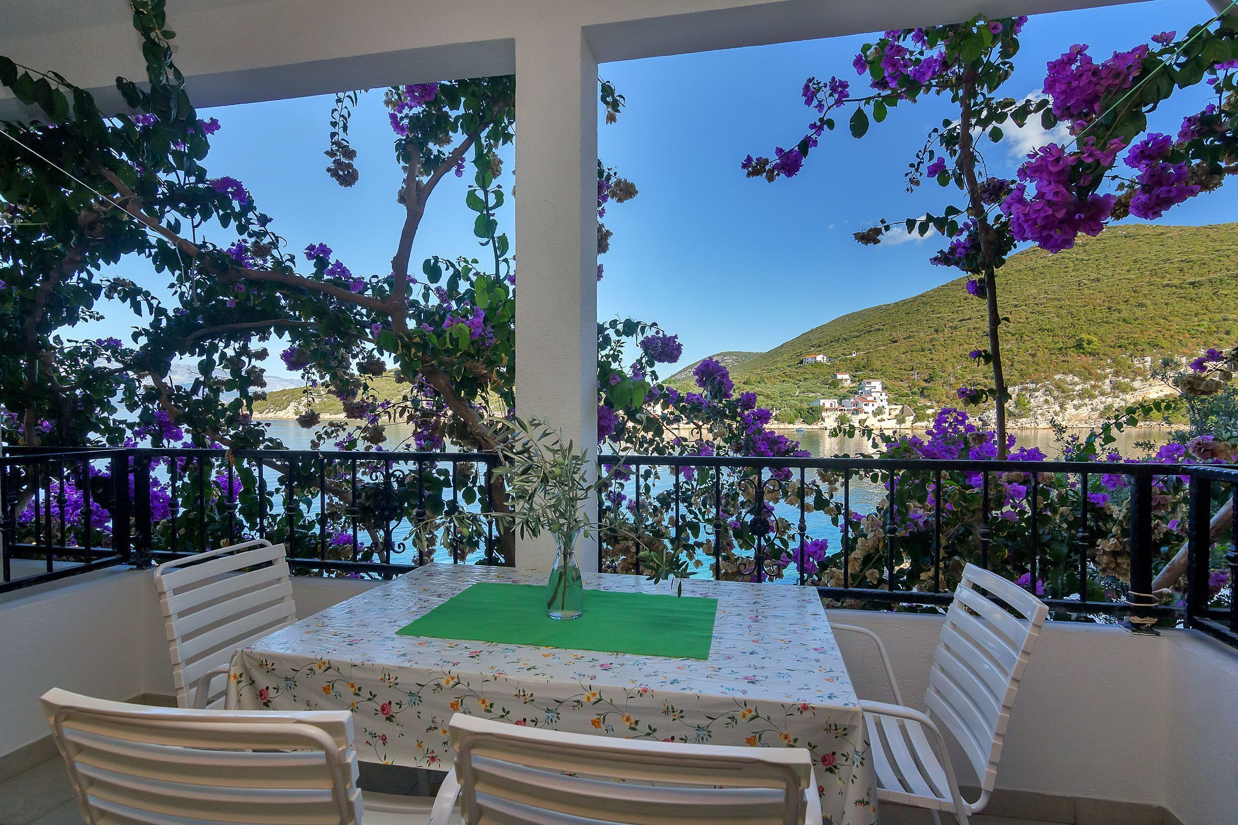 A balcony with a table and chairs, overlooking a scenic coastal view with purple flowers and a hilly landscape.