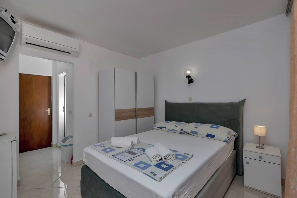 A hotel room with a bed, bedside table, nightstand, wardrobe, and an air conditioning unit.