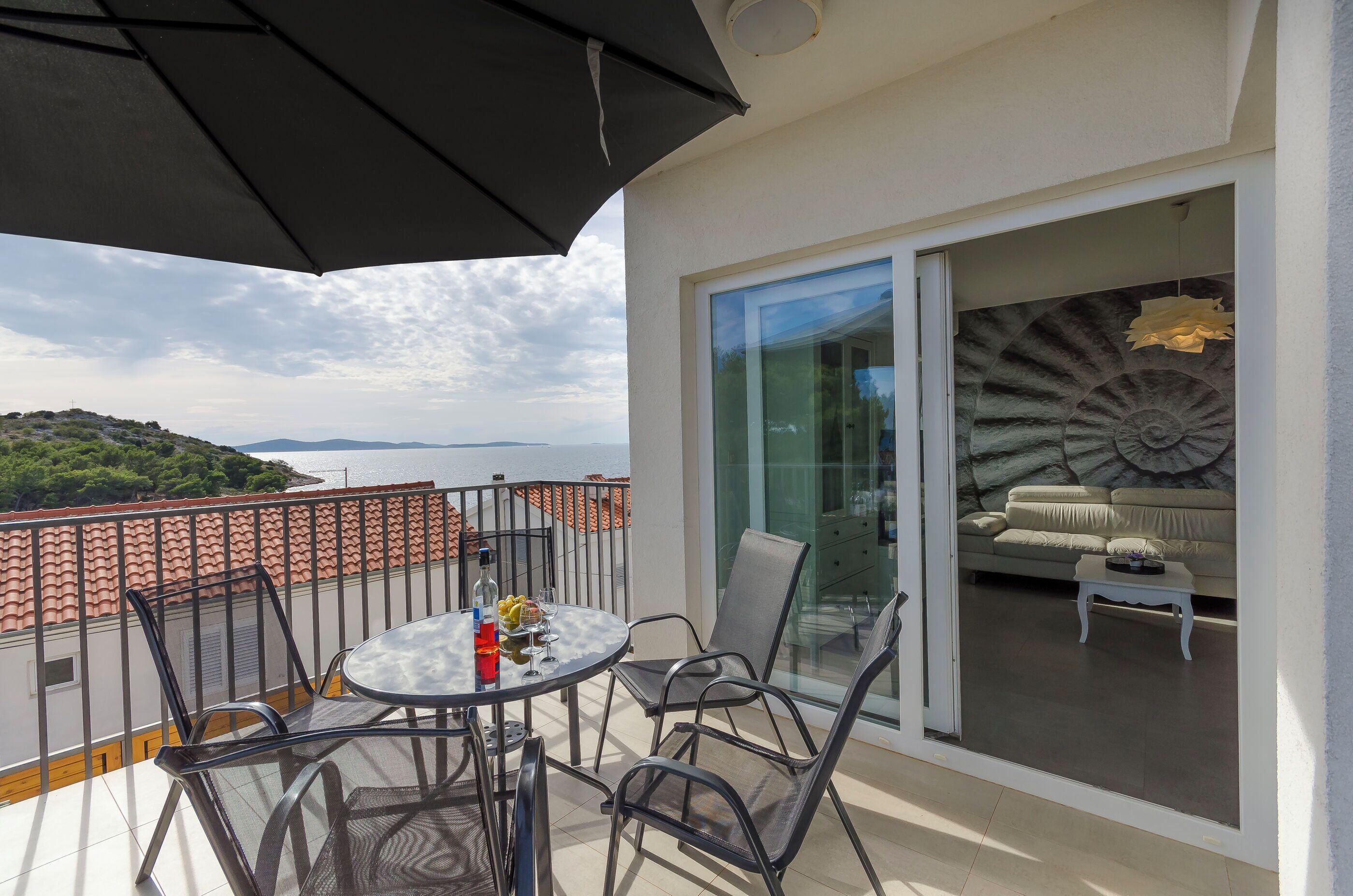 Un balcon avec une table et des chaises, une porte vitrée donnant sur un espace de vie comprenant un canapé et un élément décoratif mural.