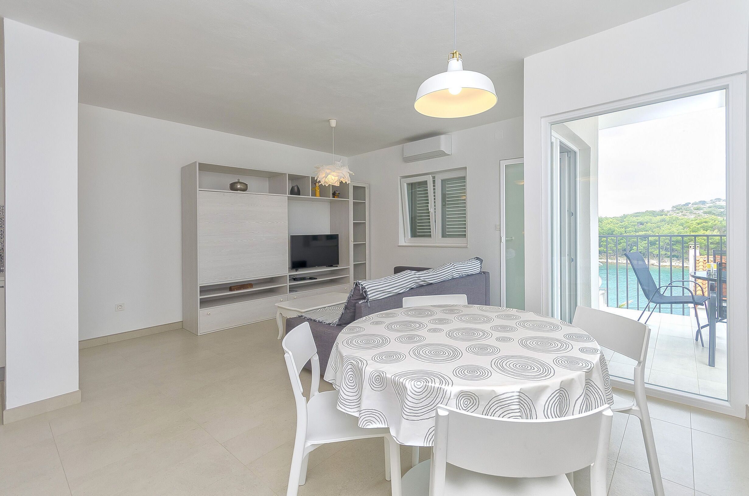 A modern dining area with a round table, white chairs, and a patterned tablecloth. There is a built-in TV shelf, a sofa with a striped blanket, and a balcony with a view of the sea.