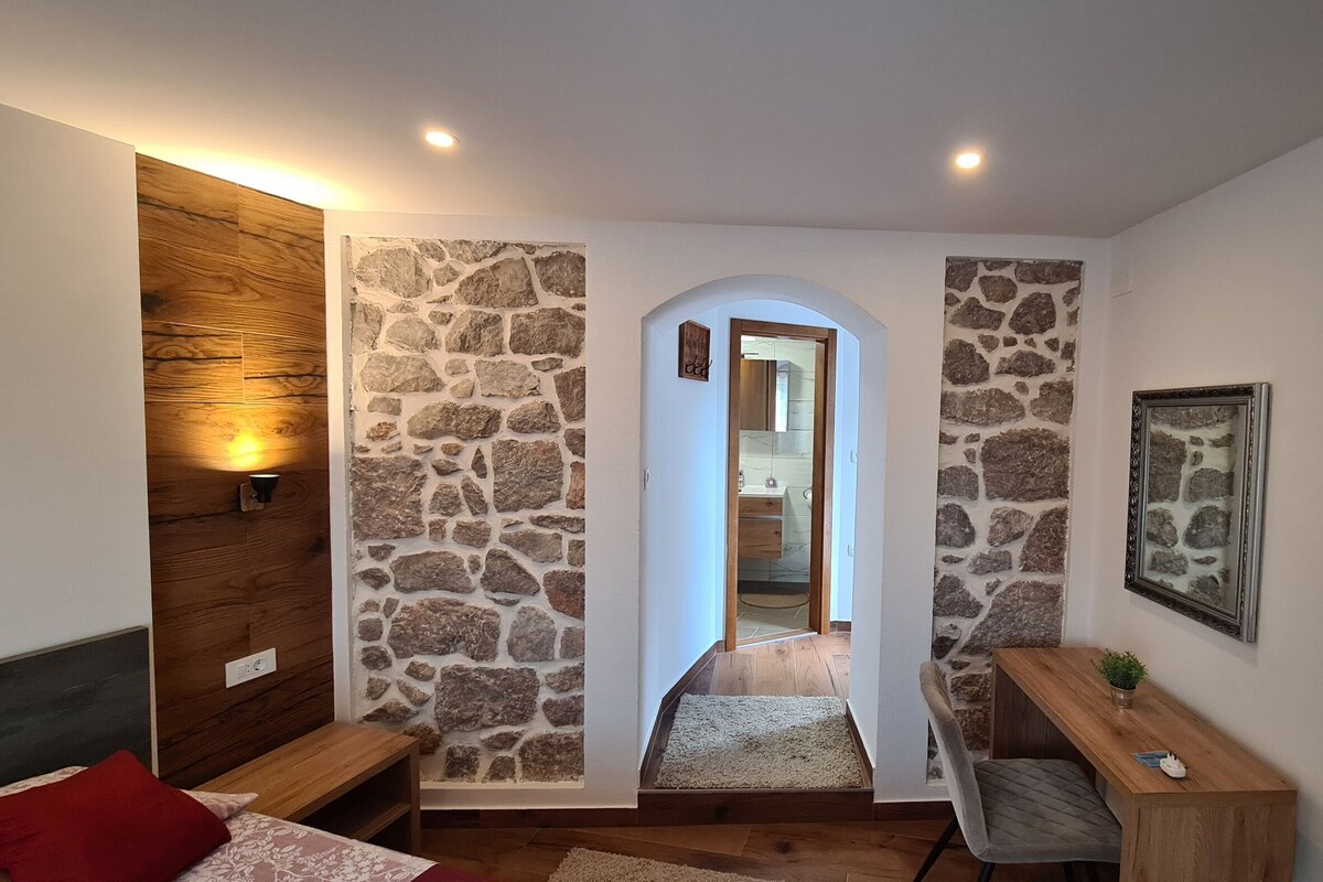 Une pièce avec un mur en pierre, une porte en bois, un bureau, une chaise, un lit et un téléviseur.