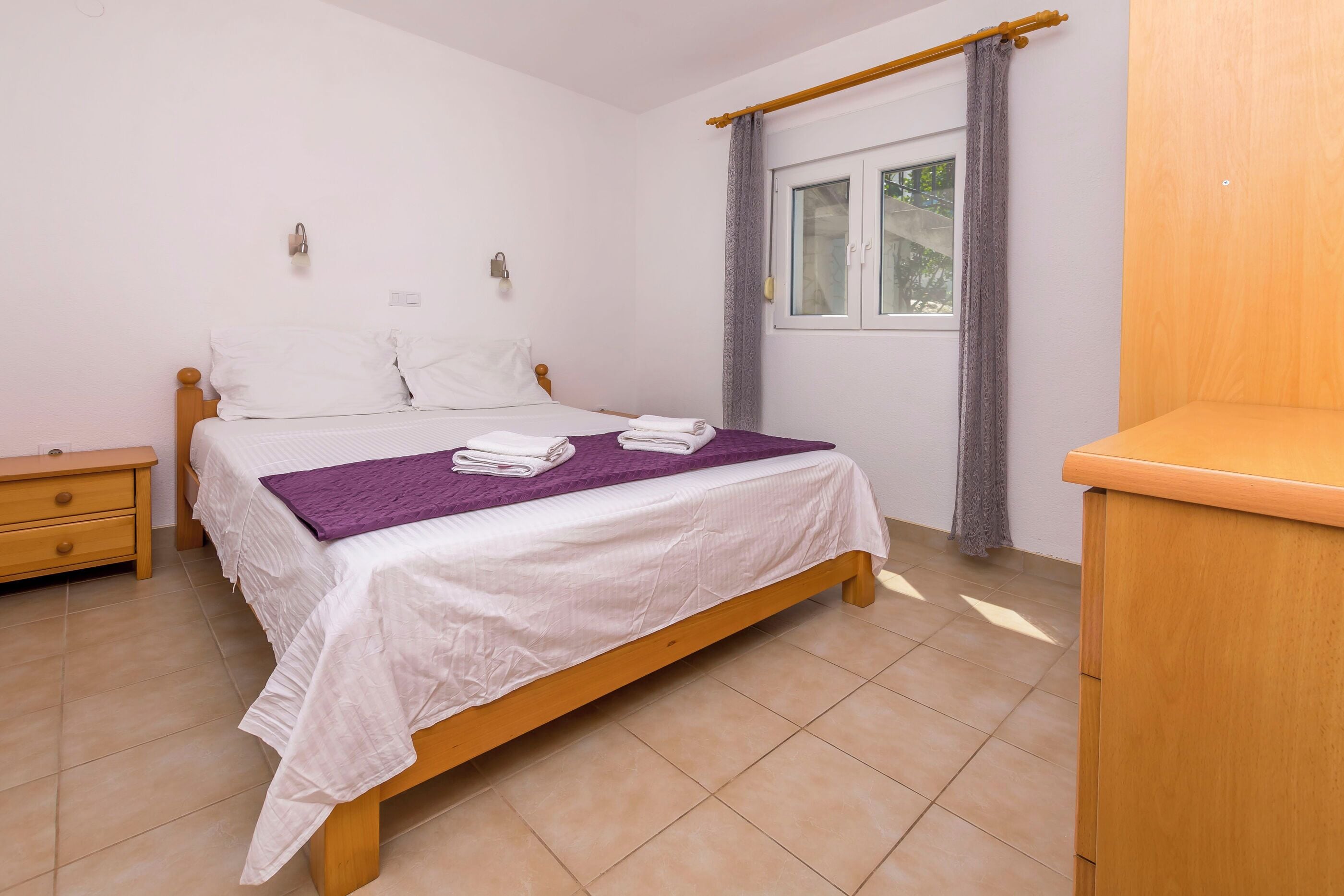 A bedroom with a bed, a nightstand, a wooden dresser, a window with curtains, and a wall-mounted light fixture.